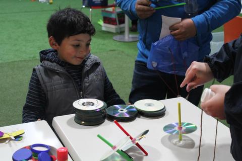 Un niño sonriente juega con discos en una mesa mientras otros adultos están involucrados en una actividad creativa.