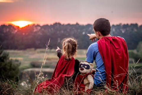 Dos niños con capas de superhéroe observan el atardecer señalando hacia el horizonte.