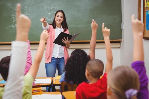 Una maestra en el aula enseña a sus estudiantes, quienes levantan la mano para participar.