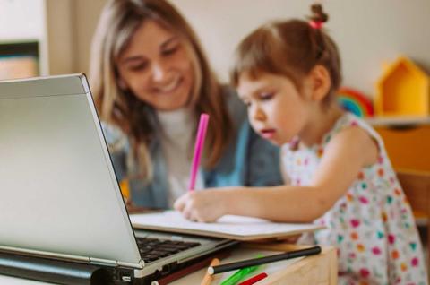 Una madre sonriente ayuda a su hija a dibujar frente a una computadora portátil en un ambiente acogedor.