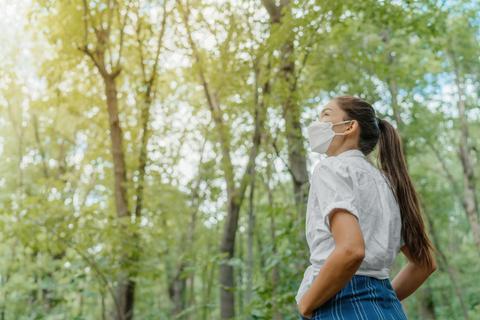 Una mujer con mascarilla observa un paisaje arbolado en un día soleado.
