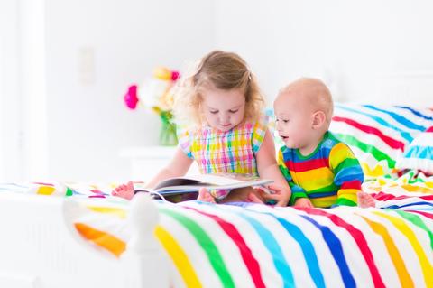 Un niño y una niña están sentados en una cama de sábanas multicolores, leyendo un libro juntos.