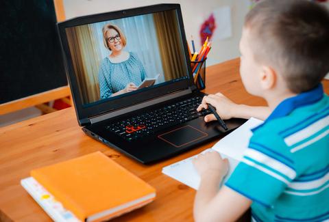 Un niño está sentado frente a un ordenador portátil, viendo a una mujer que aparece en la pantalla mientras toma notas en un cuaderno.