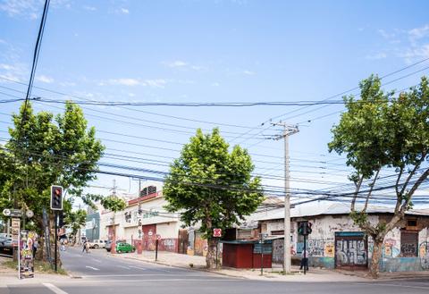 Una calle urbana con edificios y árboles, donde se pueden ver instalaciones eléctricas en el aire.