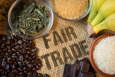 Una composición de productos como café, arroz, azúcar, té, chocolate y plátanos sobre un fondo de yute con la inscripción 'Fair Trade'.