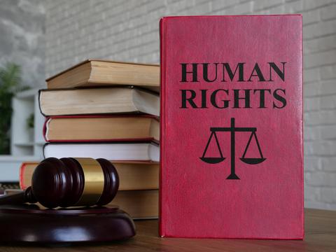 Un libro rojo titulado 'Derechos Humanos' está en primer plano junto a varios libros apilados y un martillo de juez.