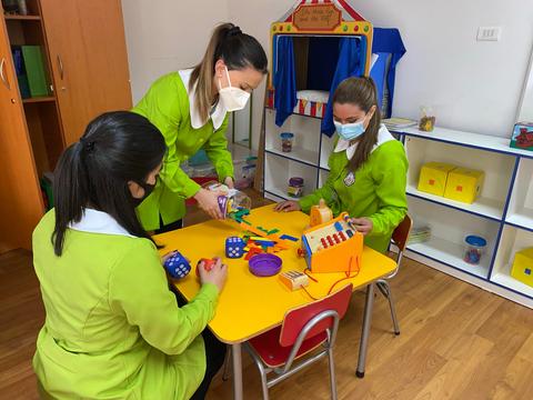 Tres personas en un aula de preescolar juegan con juguetes educativos en una mesa amarilla.