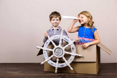 Dos niños juegan con un timón de barco y una caja de cartón como si fueran navegantes.