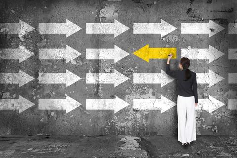 Una mujer está pintando una flecha amarilla en una pared cubierta de flechas blancas, simbolizando un enfoque diferente o un cambio de dirección.