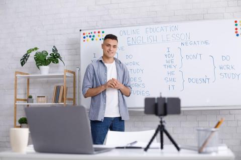 Un joven sonriente enseña una lección de inglés en un aula virtual con un pizarrón detrás.