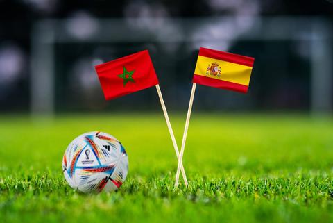 En la imagen se pueden ver dos banderas, una de Marruecos y otra de España, junto a un balón de fútbol sobre un césped verde.