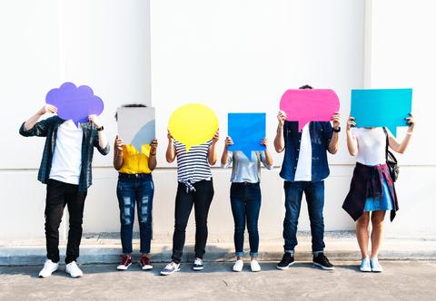 Un grupo de seis personas sostiene carteles de diferentes colores frente a sus rostros en un paisaje urbano.