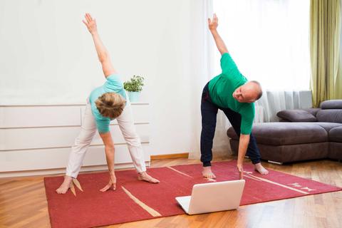 Una pareja de personas mayores practicando yoga en casa, siguiendo un video en una computadora portátil.
