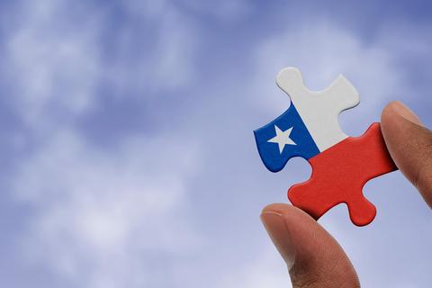 Una mano sostiene una pieza de rompecabezas con la bandera de Chile contra un fondo de cielo despejado.