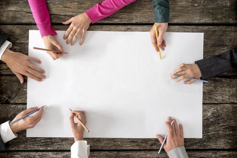 Varias manos sosteniendo lápices alrededor de una hoja en blanco sobre una mesa de madera.