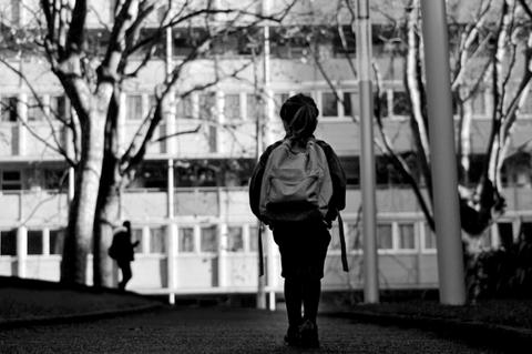 Un niño camina por un camino, con un edificio y árboles al fondo, en una escena en blanco y negro.