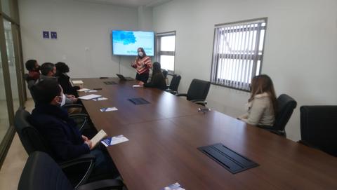 Una reunión en una sala con varias personas sentadas alrededor de una mesa, mientras una mujer presenta en una pantalla.