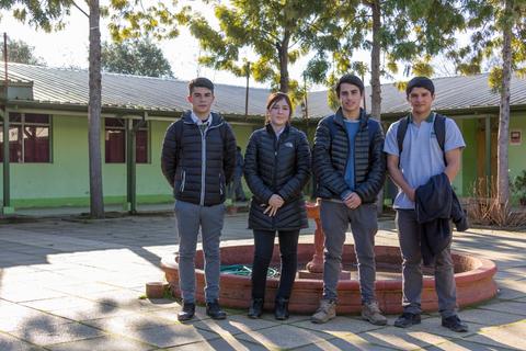 Cuatro jóvenes posan juntos en un espacio al aire libre con edificios de fondo.