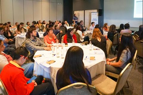 Un grupo de personas en una conferencia sentadas alrededor de mesas en un salón.
