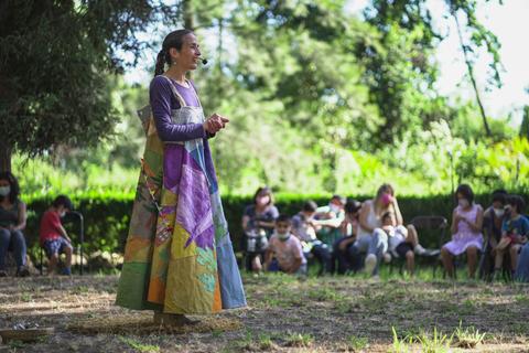 Una narradora cuenta historias al aire libre frente a un grupo de niños sentados en un círculo.