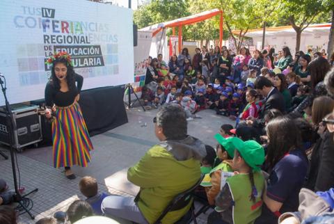 Una mujer vestida con una falda de colores presenta un espectáculo ante un grupo de niños y adultos en un evento al aire libre.