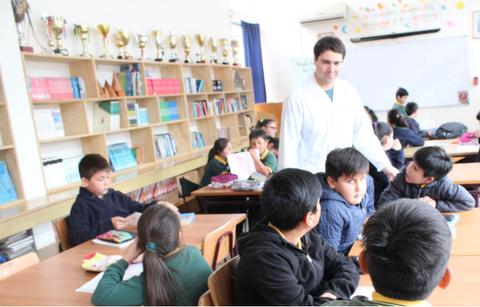 Un profesor interactúa con estudiantes en un aula mientras otros trabajan en sus tareas.