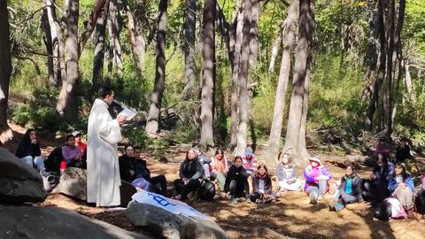 Un grupo de personas escucha atentamente a un orador en un entorno natural, rodeado de árboles.