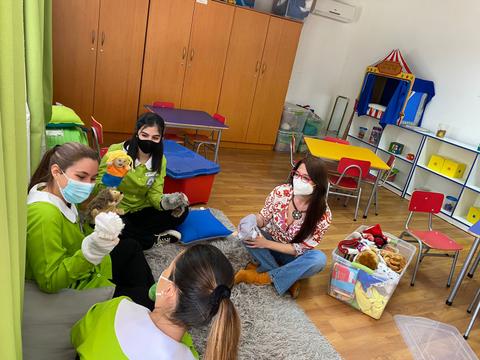 Un grupo de cuatro personas se encuentra en un aula de educación infantil, interactuando con títeres y juguetes.