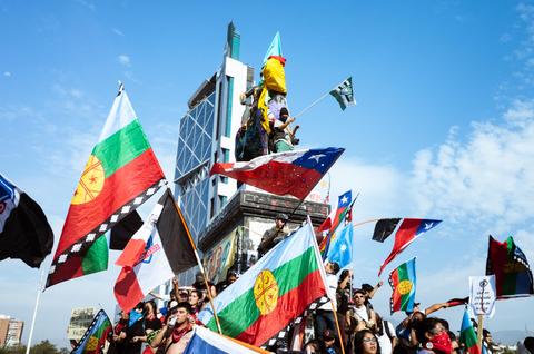 Un grupo de personas sostiene banderas en una manifestación frente a un edificio en un día soleado.