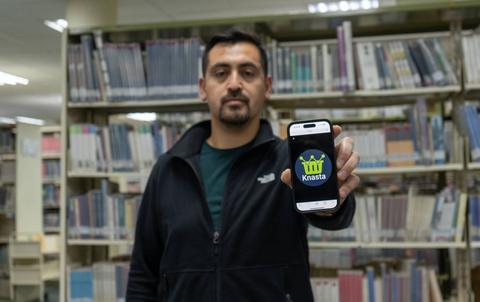 Un hombre sostiene un teléfono móvil con la aplicación Knasta frente a una estantería de libros en una biblioteca.