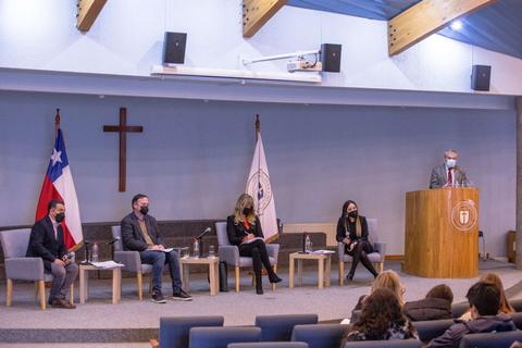 Una mesa de discusión en un auditorio con panelistas, banderas y un cruz al fondo.
