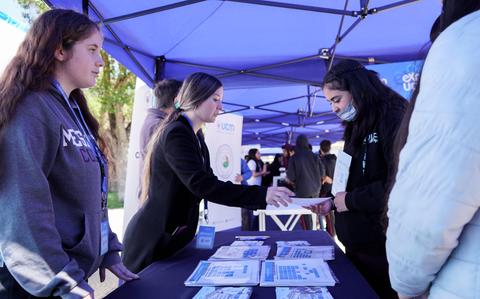 Un grupo de personas interactúa en un evento bajo carpas, mientras que una de ellas entrega un folleto.