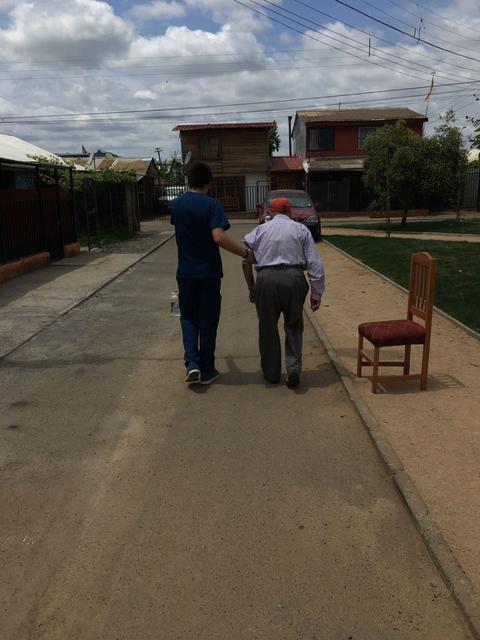 Un joven acompaña a un anciano por una calle tranquila, mientras una silla vacía se encuentra al borde de la acera.