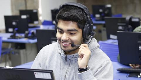 Un joven sonriente usa auriculares y un micrófono mientras trabaja en una computadora en un aula.