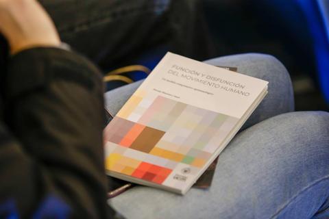 Un libro titulado 'Función y Disfunción del Movimiento Humano' reposando sobre las piernas de una persona.