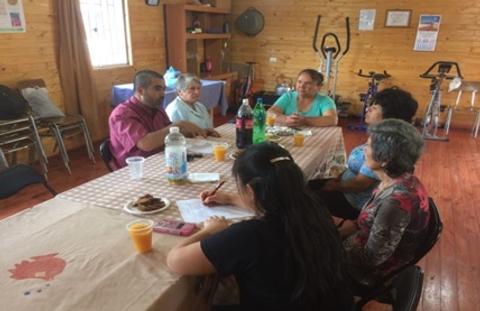 Un grupo de personas mayores se reúne en una sala conversando y compartiendo alimentos y bebidas.
