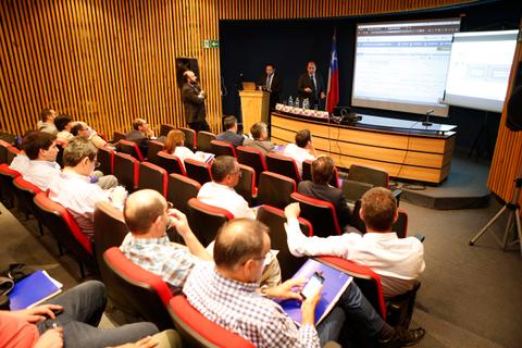 La imagen muestra una sala de conferencias con un grupo de personas sentadas y un presentador en el escenario.