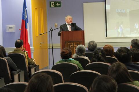 Un ponente habla frente a un público en una sala con una bandera chilena en el fondo.