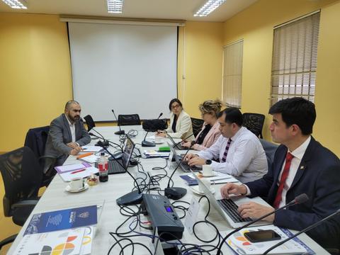 Una reunión de trabajo en una sala con seis personas sentadas alrededor de una mesa con computadoras portátiles y documentos.
