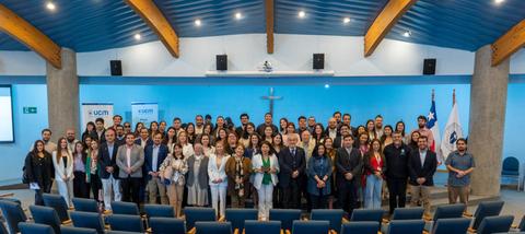 Un grupo numeroso de personas posando en un salón con un fondo azul y banderas.