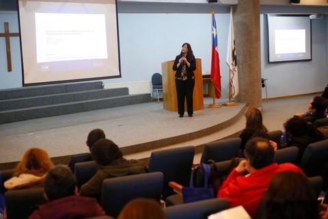 Una presentadora habla frente a una audiencia en un salón con banderas al fondo.