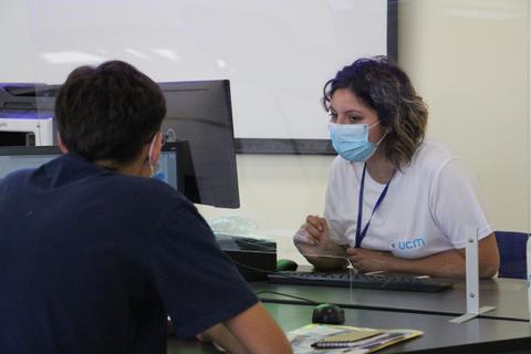 Una mujer con mascarilla atiende a un joven en una mesa de trabajo.