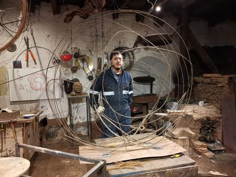 Un hombre se encuentra en un taller rodeado de estructuras de alambre y herramientas de trabajo.