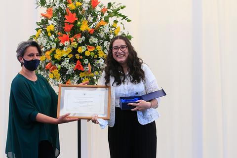 Dos mujeres sonríen mientras sostienen un certificado frente a una floristería colorida.