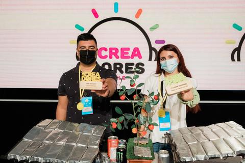 Dos personas están en un evento sosteniendo premios y con productos sobre una mesa.