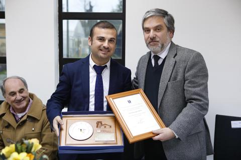 Dos hombres sonríen mientras sostienen un premio y un certificado en un evento formal.