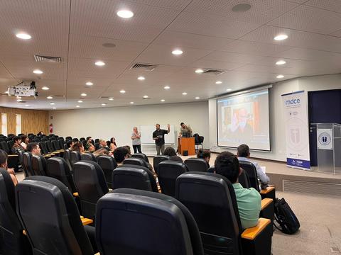La imagen muestra a un grupo de personas escuchando una presentación en un auditorio.