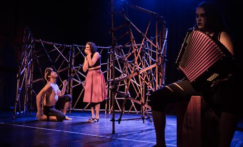 Una actuación teatral en un escenario con tres actores, donde uno toca el acordeón, mientras los otros interactúan en un ambiente dramático.