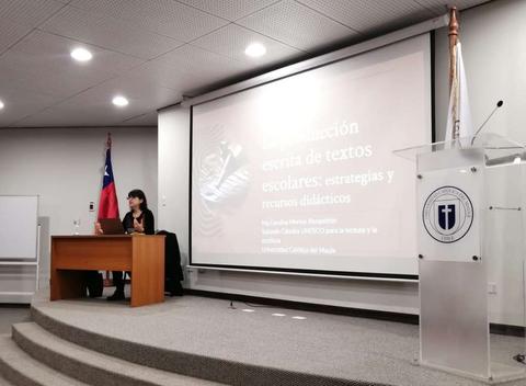 Una mujer presenta una charla sobre la escritura de textos escolares en un aula con una pantalla grande.