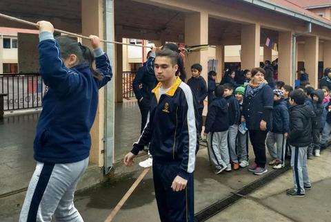 Un grupo de niños está haciendo fila en un patio escolar mientras uno de ellos intenta saltar un palo sostenido por otro.